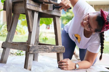 Kadın bahçedeki sandalyeyi boyamak ve restore etmek için öne eğiliyor.