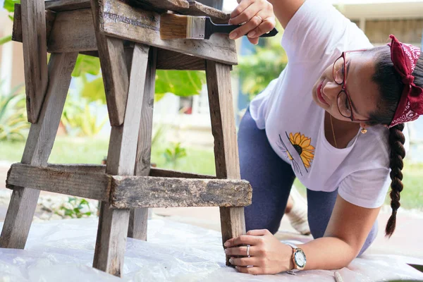 Kadın bahçedeki sandalyeyi boyamak ve restore etmek için öne eğiliyor.