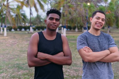 Spor giyimli iki arkadaş, parkta durmuş, kollarını kavuşturup gülümsüyorlar..