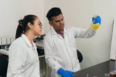 Ciddi laboratuvar çalışanları gözlerinin önünde bir balon tutuyor ve içindekileri inceliyor. Koruyucu eldiven, gözlük ve beyaz önlük giyen bilim adamları. Laboratuvar ve kimya kavramı