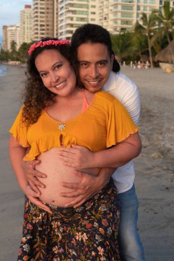 Latin koca sahilde hamile karısına sarılıyor.