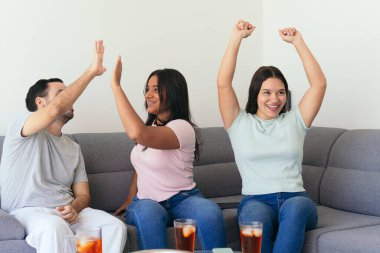 Arkadaşlar galibiyet golünü atarken beşlik çakıyor. Futbol hayranları kanepede otururken içki ve atıştırmalıkların tadını çıkarıyorlar. Evde çok eğleniyorlar..