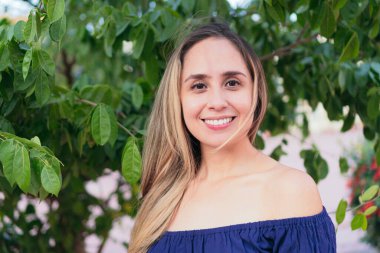 Güneşli bir günde şehir parkında dikilirken günlük giysiler içinde gülümseyen ve kameraya bakan bir kadın..