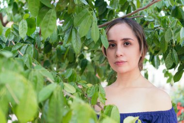 Genç bir kadın, yemyeşil bir parkta çeşitli bitkilerle çevriliyken poz veriyor.