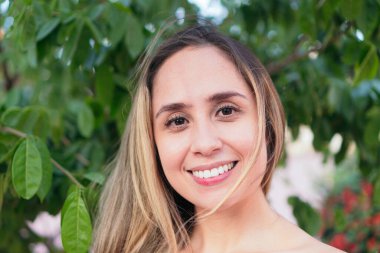 Bir şehir parkında dururken gülümseyen ve yan bakan Latin kadın..