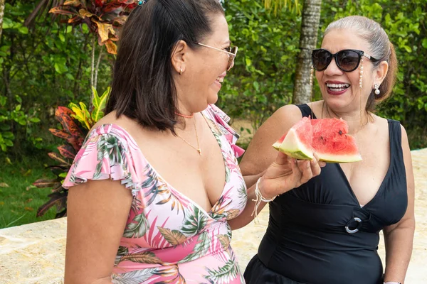 Older women in bikini sitting by the pool and eating watermelon. Happy friends having a summer picnic outdoors.