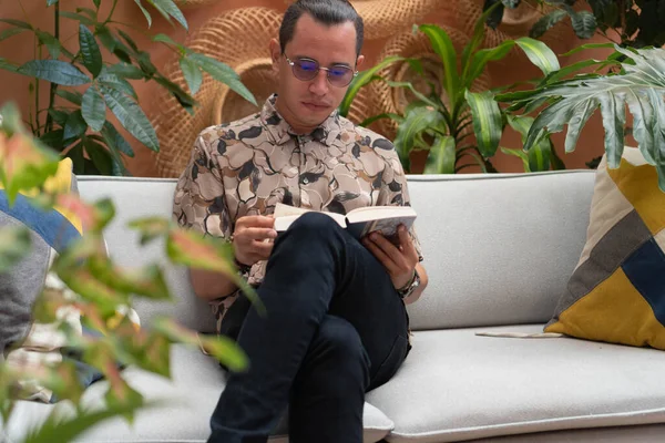 Latin Man sitting in an armchair reading a book. Young man reading a book in his cozy apartment.