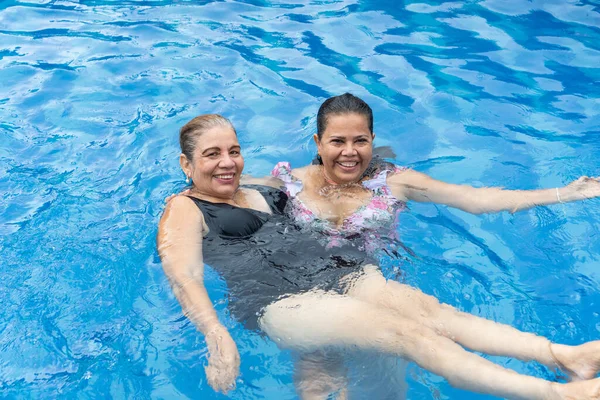 Two mature women with wet hair and smiling in bikinis swimming in the pool.