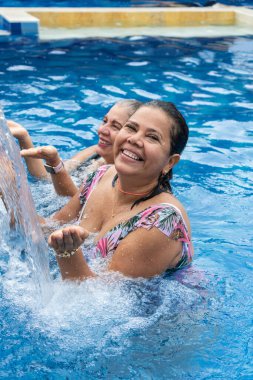 Smiling mature women with wet hair and bikini under the water jet in the swimming pool.