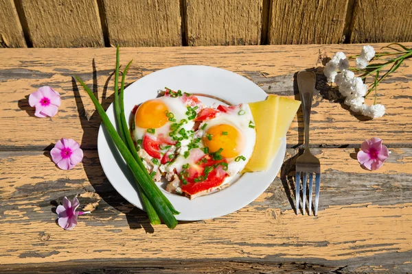 Flores cor-de-rosa, um prato com ovos, cebolas verdes e tomates na velha mesa de madeira — Fotografia de Stock