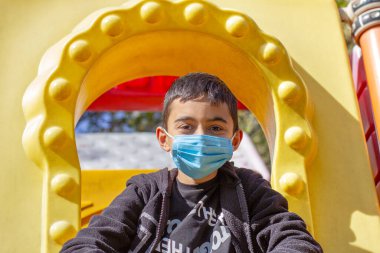 Parktaki maskeli çocuk. Siyah kapüşonlu ve mavi maskeli esmer çocuk parkta oynuyor. Ön manzara. Seçici odak