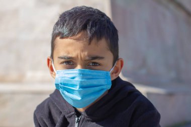 Sıkılmış, hayal kırıklığına uğramış çocuk maskesi takıyor. Siyah gömlekli ve mavi maskeli esmer çocuk. Seçici odak