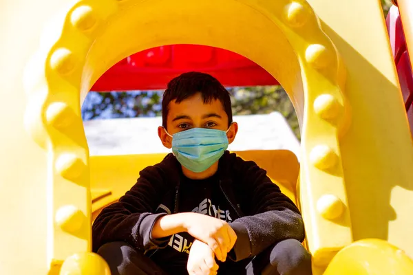 Parktaki maskeli çocuk. Siyah kapüşonlu ve mavi maskeli esmer çocuk parkta oynuyor. Ön manzara. Seçici odak