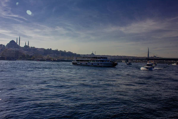 Eminonu, İstanbul, Türkiye-Ekim-Cumartesi-2021: Denizde küçük bir tekne hareket ediyor. Arkasında büyük bir yolcu feribotu var. Süleyman Camii ve Altın Boynuz Metro Köprüsü manzarası
