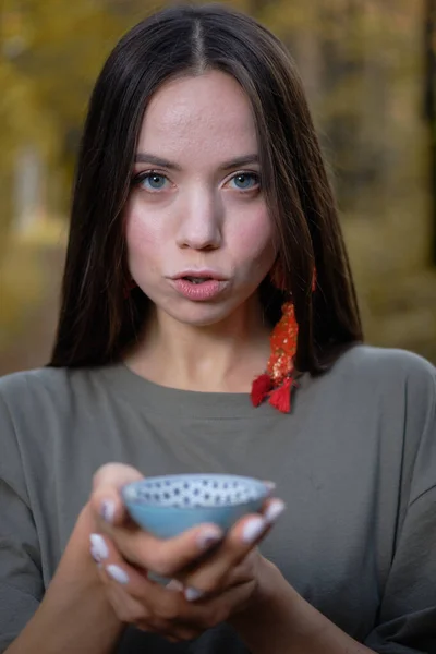 Jeune belle fille assise sur le sol en vêtements modernes dans la forêt jaune d'automne et tenant une cérémonie du thé Photo De Stock