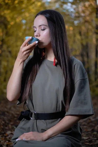 Młoda piękna dziewczyna siedzi na ziemi w nowoczesnych ubraniach w jesiennym żółtym lesie i gospodarstwa ceremonii herbaty Obrazy Stockowe bez tantiem