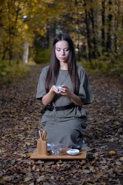 Jeune belle fille assise sur le sol en vêtements modernes dans la forêt jaune d'automne et tenant une cérémonie du thé Images De Stock Libres De Droits