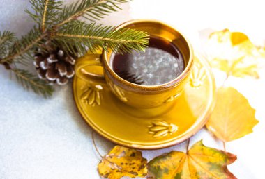 Sonbahar çay partisi. Gövde üzerindeki küçük sarı seramik kahve fincanı Noel ağacı dalının yanındaki beyaz bir masanın üzerinde duruyor, köknar konisi, düşmüş yapraklar. Güzel doğa kompozisyonu. Mesaj için yer. Sıcak bir sabah.