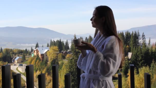 魅力的なブルネットの若い女性は きれいな朝の空気中で呼吸し バスローブでお茶を保持するかわいい女の子 バルコニーに立ってコーヒーカップ 森林の素晴らしい景色 スパレクリエーション屋外 — ストック動画