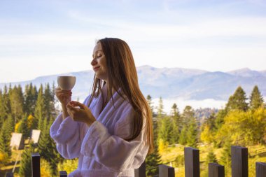 Bornozlu güzel esmer kız elinde çay fincanı, sabahın köründe balkonda duran kahve, temiz havayı zevkle soluyor. Tepeler, ağaçlar, dağlar. Spa eğlencesi. Doğa dışarıda..