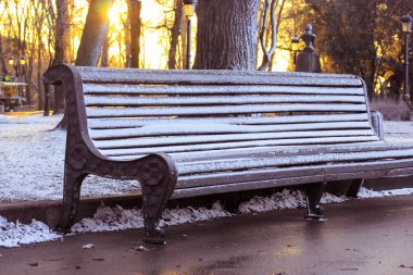 Gün batımında şehir parkında karla kaplı ahşap bir bank. Sarı parlak gökyüzü ve çıplak ağaçların arkasında güneş ışığı. Soğuk bir kış gününde ilk kar yağacak. Ocak, Aralık, Şubat. Dışarıda dinlenecek bir yer..
