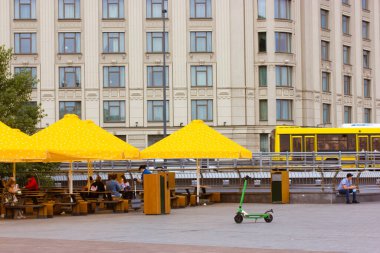 Kyiv, Ukrayna. 8 Temmuz 2021. Kafe terası, sarı şemsiyeli bistro. Açık masalar, yemek servisi için sandalyeler. Büyük şehir hayatı. Sokaktaki insanlar. Scooter kaldırıma park etmiş.