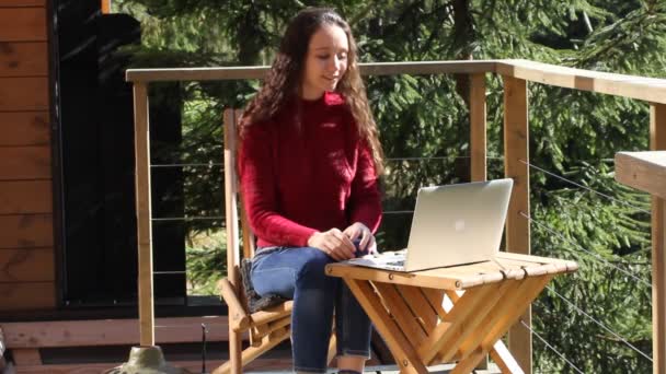 バルコニーテラスの木製の椅子に座ってラップトップでビデオ通話を介して女の子チャット オンライン通信 若い女性は 自然の中で晴れた秋の日にリラックスしてお茶を飲む 森の中の小屋森の家 — ストック動画
