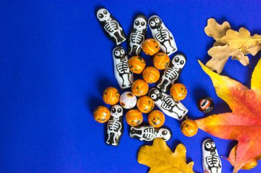 Siyah beyaz iskelet şekerler, portakallı balkabağı, düşen sonbahar akçaağacı, mavi arka planda yer alan meşe yaprakları. Cadılar Bayramı konsepti. 31 Ekim 'de bir partiye davet.