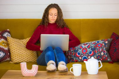 Güzel esmer kız renkli dekoratif yastıkların arasında dizüstü bilgisayarla kanepede oturuyor. Uzaktan çalışan, çalışan, sonbahar kışında internetten alışveriş yapan genç bir kadın. Çay fincanlı ahşap masa, çaydanlık