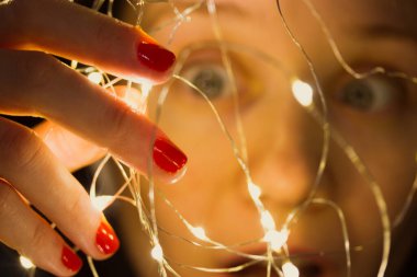 Şaşırmış bir kız elinde parlak ışıklarla karışık bir çelenk tutuyor. 2022 yılbaşı arifesi için Noel ağacını süslemeye hazırlanan kadın. Kablo tutan turuncu-kırmızı manikürlü ince parmaklar..