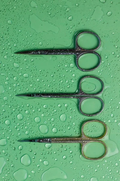 Sharp titanium manicure scissors for trimming cuticles on green background with water drops flatly with copy space. Metal tools for work of manicurist in beauty salon. Nail pedicure, body care concept