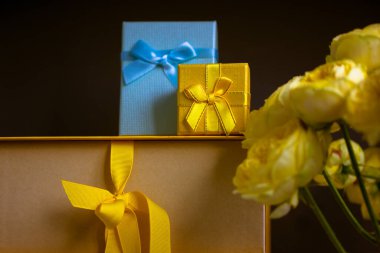 Koyu gri arka planda bir sürü sarı mavi hediye kutusu, sarı Austin gül buketi. Hediyelerle dolu tebrik kartı, doğum günü hediyeleri, Anneler Günü, yıldönümü. Alışveriş, satış, hediye..