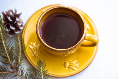 Bir fincan tabağının üzerindeki küçük sarı seramik kahve fincanı beyaz bir masa arkasında duruyor. Noel ağacı dalı ve köknar konisi. Güzel minimalist doğa kompozisyonu. Mesaj için yer. Rahat bir ev. Kış ikilisi.