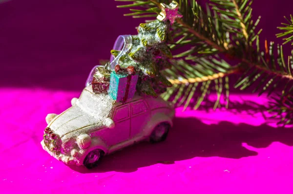 White toy car with green pine Christmas tree with a gift box on a roof on a bright fuchsia pink background. Happy 2022 New Years Eve. Greeting card copy space. Gifts delivery. Contrasting shades. — Fotografia de Stock
