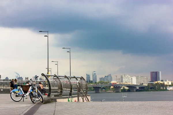 Un paseo marítimo en un día nublado con nubes azules ante la lluvia. Una bicicleta de alquiler se encuentra en una acera. Paisaje urbano. Las farolas en el fondo de un cielo sombrío. Un río, edificios. — Foto de Stock