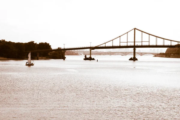 Kiev, Ukraine. 1er juillet 2021. Pont piétonnier au-dessus d'une rivière Dniepr à Kiev. Beau paysage carte postale crépuscule vue du soir d'un voilier flottant au bord de l'eau et des ponts sur un horizon. Tonification brune. — Photo