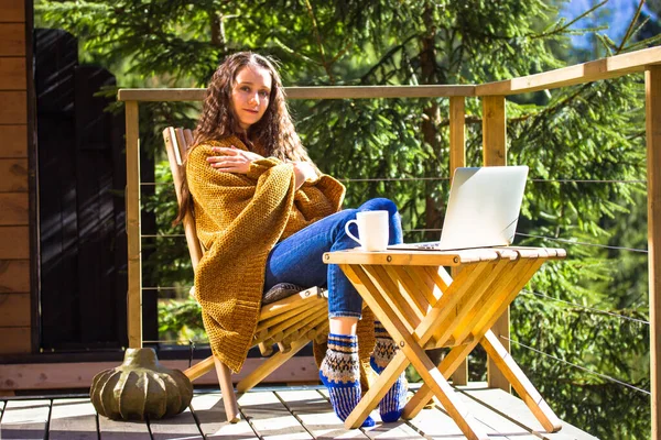 Κορίτσι κάθεται στο μπαλκόνι τυλιγμένο σε καρό, απολαμβάνοντας την ημέρα του φθινοπώρου. Εργασία σε φορητό υπολογιστή — Φωτογραφία Αρχείου