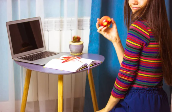 Όμορφη μαθήτρια κορίτσι σε πολύχρωμο πουλόβερ με φορητό υπολογιστή ατμόσφαιρα σπίτι τρώει μήλο. — Φωτογραφία Αρχείου