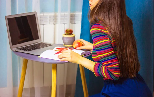 Όμορφη μελαχρινή μελαχρινή κοπέλα σε πολύχρωμο ριγέ πουλόβερ με φορητό υπολογιστή σε ένα τραπέζι στο σπίτι ατμόσφαιρα. Γυναίκα φοιτήτρια με κόκκινο μήλο σπουδάζει στο σπίτι, εργάζεται εξ αποστάσεως, κρατώντας σημειώσεις σε ένα σημειωματάριο. — Φωτογραφία Αρχείου