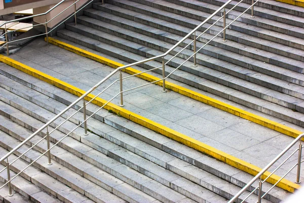 A wide, modern stone staircase with yellow markings for blind people and metal handrails for a convenience of pedestrians. The steps go downhill view from above. A deserted street. City infrastructure — Stock Photo, Image