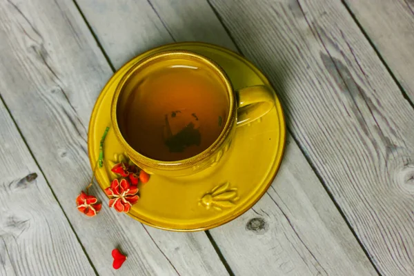 Taza de cerámica amarilla con té negro en platillo sobre mesa de madera gris. Arbusto del huso, Euonymus europaeus, Pfaffenhutchen pétalos de flores rojas. Pétalo en forma de corazón. Composición plana de naturaleza muerta. —  Fotos de Stock