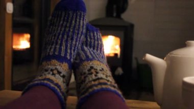 Akşam evde şöminenin yanında dinlenen genç bir kadın, şöminenin yanında ayaklarını yünlü mavi örgü çoraplarla ısıtıyor. Huzur, uyum konsepti. İçeriden sıcak ve güzel bir ev. Sıcak bir yer..