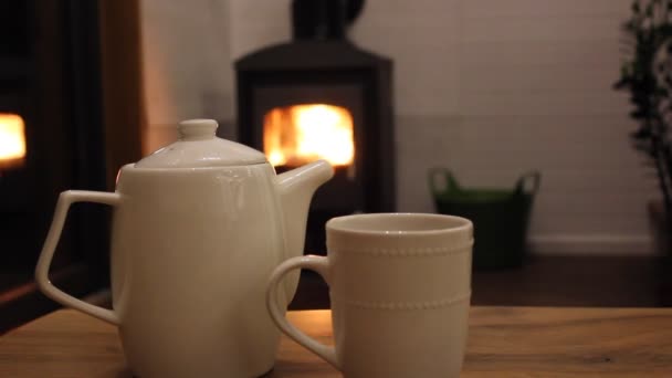 Kahve içmek için beyaz seramik çaydanlık ve fincan, ahşap bir masada çay standı. Şöminede ateş içinde odun var. Sıcak, sade bir atmosfer. Sonbahar akşamı. Modern iç kısımlarda sıcak tutun.. — Stok video
