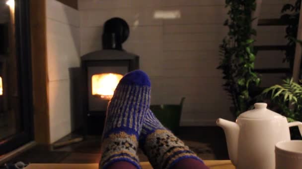 Eine junge Frau entspannt sich abends zu Hause am Kamin und wärmt ihre Füße in wollblauen Stricksocken am Feuer. Ruhe, Harmonie. Warme, süße Hauseinrichtung von innen. Gemütlicher Ort. — Stockvideo
