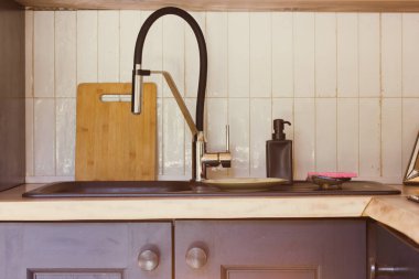 Seramik lavabo, siyah su musluğu, beyaz seramik fayanslara karşı su musluğu. Minimalist tarzda modern ahşap mutfağın içi. Tahta kesme tahtası, sıvı sabun için siyah kap. Mutfak eşyaları..