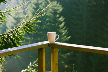 Beyaz seramik çay, kahve içeceği tahta balkonda duruyor yeşil kozalaklı orman zeminine karşı korkuluklar, uzun çam ağaçları, ormandaki ilk ağaçlar. Dağlarda kahvaltı yaparız. Güneş ışığındaki ladin dalı.