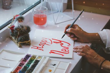 Sanatçı, kırmızı boyayla mutlu yıllar, beyaz kağıt üzerine fırça çiziyor. Şenlik arka planı. Ressam, 2022 Noel 'i için karlı bir günde DIY el yapımı kartpostal hazırlıyor. Oyuncak Noel Baba Maddesi.
