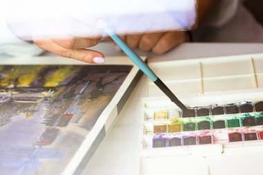 Profesyonel sanatçı elinde mavi bir fırçayla suluboya renkli resimlerle güzel bir tablo çiziyor. Kadın bir başyapıt yaratma sürecinde. Çizim için bir araç tutuyor.