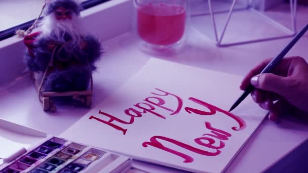 Artist draws inscription Happy New Year with red paint, brush on white sheet of paper. Painter creates DIY hand made postcard for Christmas 2022. Toy Santa Clause. The trending Pantone color Very Peri — Vídeo de Stock