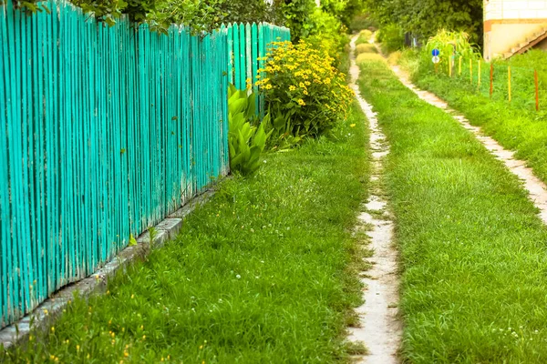 Güzel, kıvrımlı bir patika, yeşil çimlerle kaplı toprak bir yol uzak bir mesafeye gidiyor. Banliyö bölgesinde yeşil ahşap çitler. Yol, patika, yol, yol, yaz güneşli bir günde yürüyüş yolu..